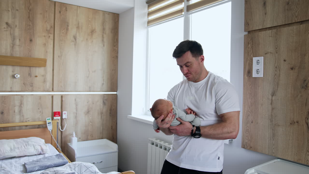 Handsome broad-shouldered Caucasian man holding a tiny baby. New-made father with a newborn in a hospital ward.
