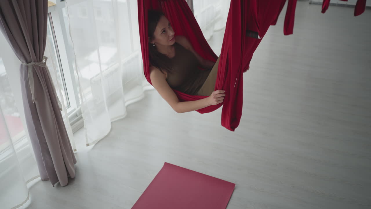 Bright yoga studio with floor to ceiling windows and sheer curtains as fitness expert lies peacefully in red silk hammock swinging gently, conveying calm balance wellness and serene flow practice