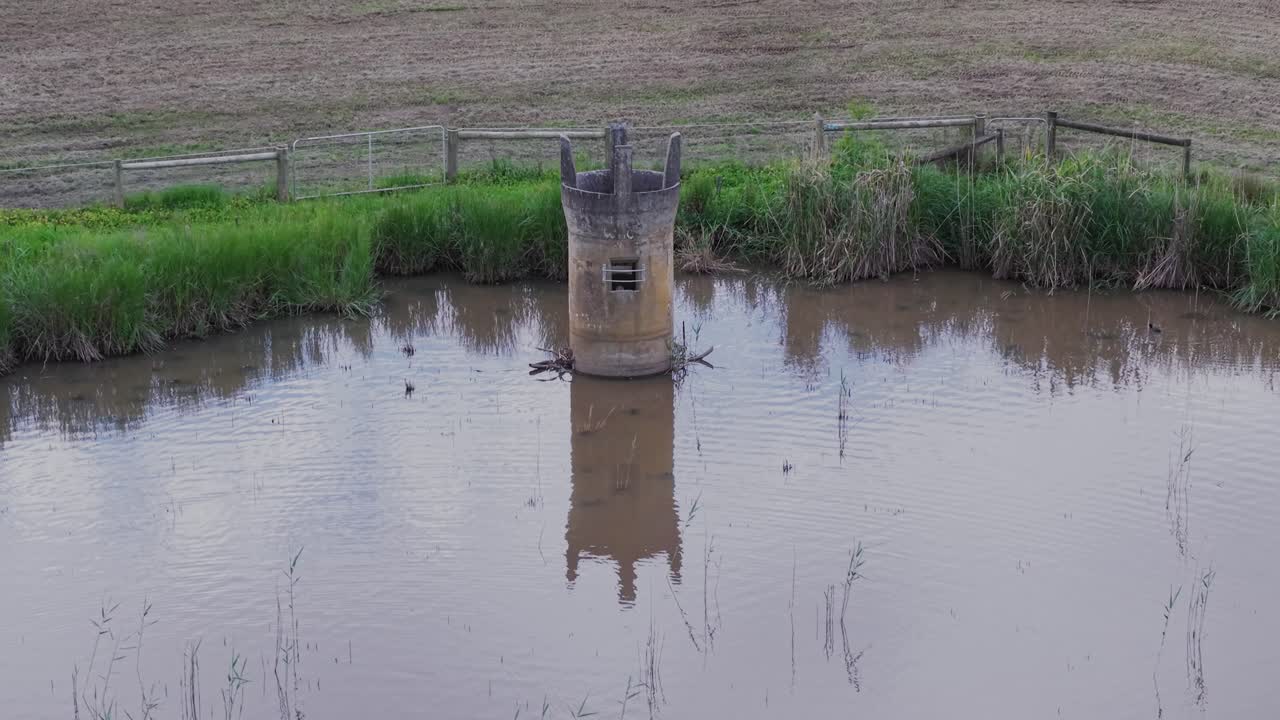 Aerial footage captures a tranquil stone tower in the middle of a serene pond, surrounded by lush greenery and fields. The calm water reflects the tower, creating a peaceful scene