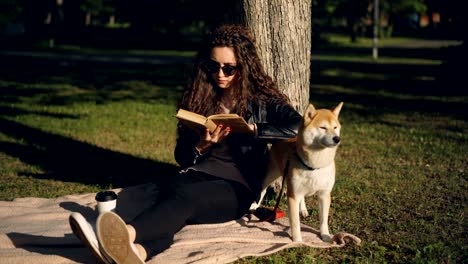 beautiful young woman is reading book sitting on blanket under tree in park and smiling while her pedigree dog is standing near her enjoying nature.