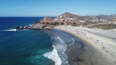 Aerial-view-over-a-beautiful-beach-overlooking-the-waves-of-the-blue-sea-with-a-view-of-beachgoers-during-their-summer-vacation-and-a-hot-dry-landscape-on-a-sunny-day-at-cerritos-beach,-mexico