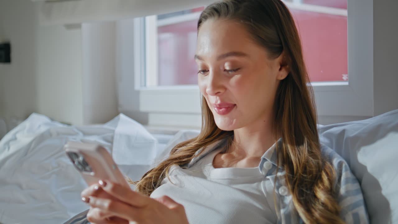 Smiling lady looking cellphone lying cozy white bedding closeup. Woman browsing