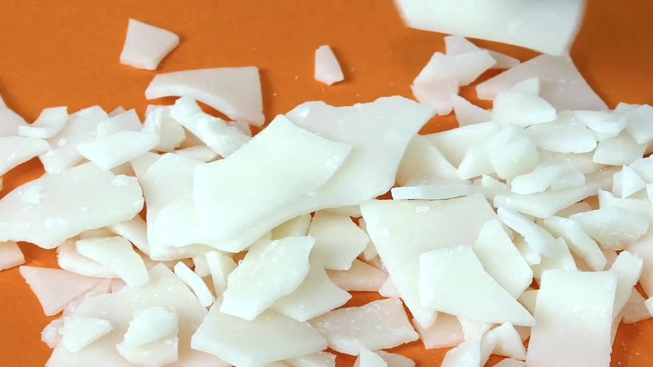 Chercheur avec des gants de protection manipulant des flocons de soude caustique sur un fond orange dans un laboratoire.