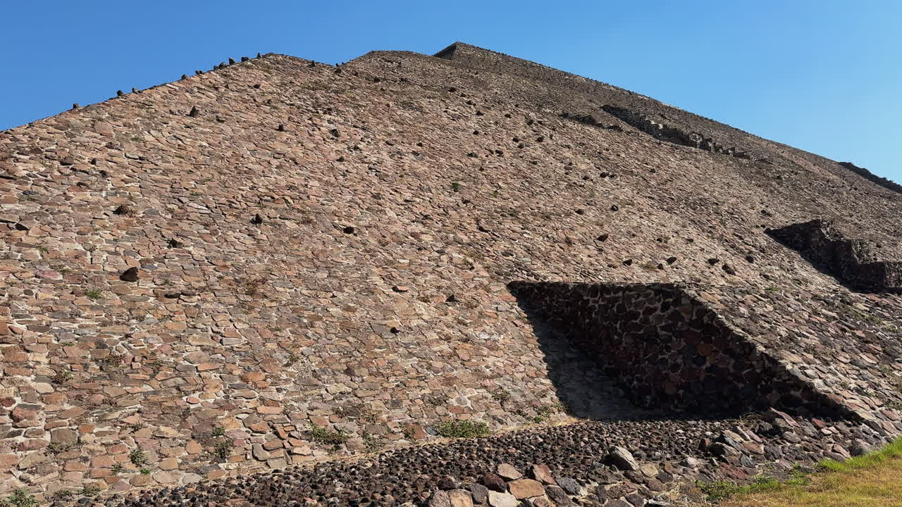 Panoramica a sinistra lungo il pendio in pietra della Piramide del Sole di Teotihuacan con nicchie in ombra e erba secca in primo piano sotto un cielo azzurro limpido, un filmato B-roll ideale per la storia di viaggio del Messico.
