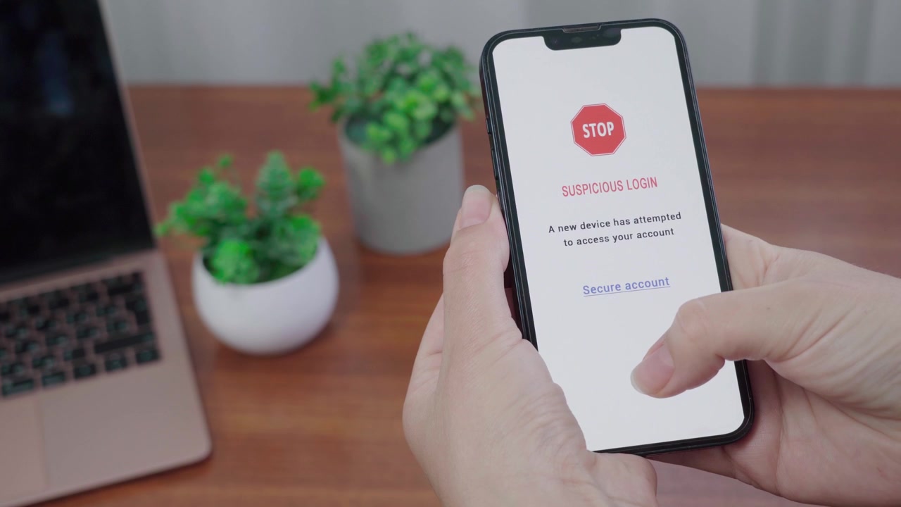 Avviso di blocco accesso sospetto appare su uno schermo informativo mobile con IA mentre un pollice si avvicina al link 'Proteggi account' su una scrivania