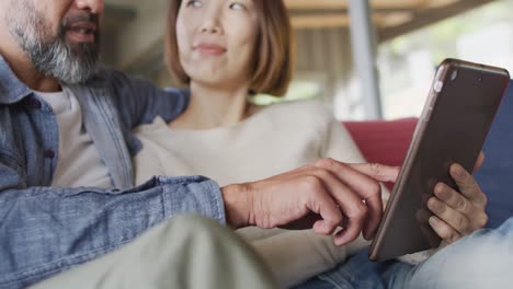 Happy-diverse-couple-sitting-in-living-room-using-tablet-and-talking