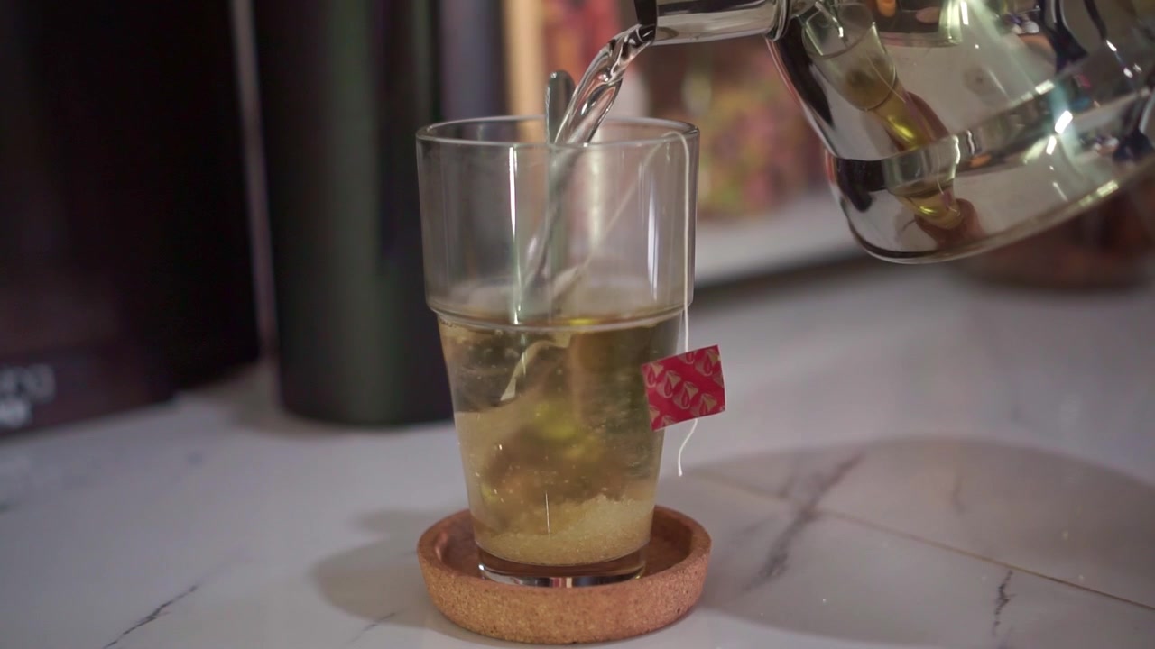 Acqua calda che si versa nel bicchiere con bustina di tè e zucchero, primo piano, preparazione casalinga del tè, sfondo bancone della cucina, bevanda
