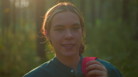 young woman in green shirt with hair tied back standing in forest, adjusting red backpack with a warm, gentle smile, sunlight filters through trees, illuminating her hair with a radiant