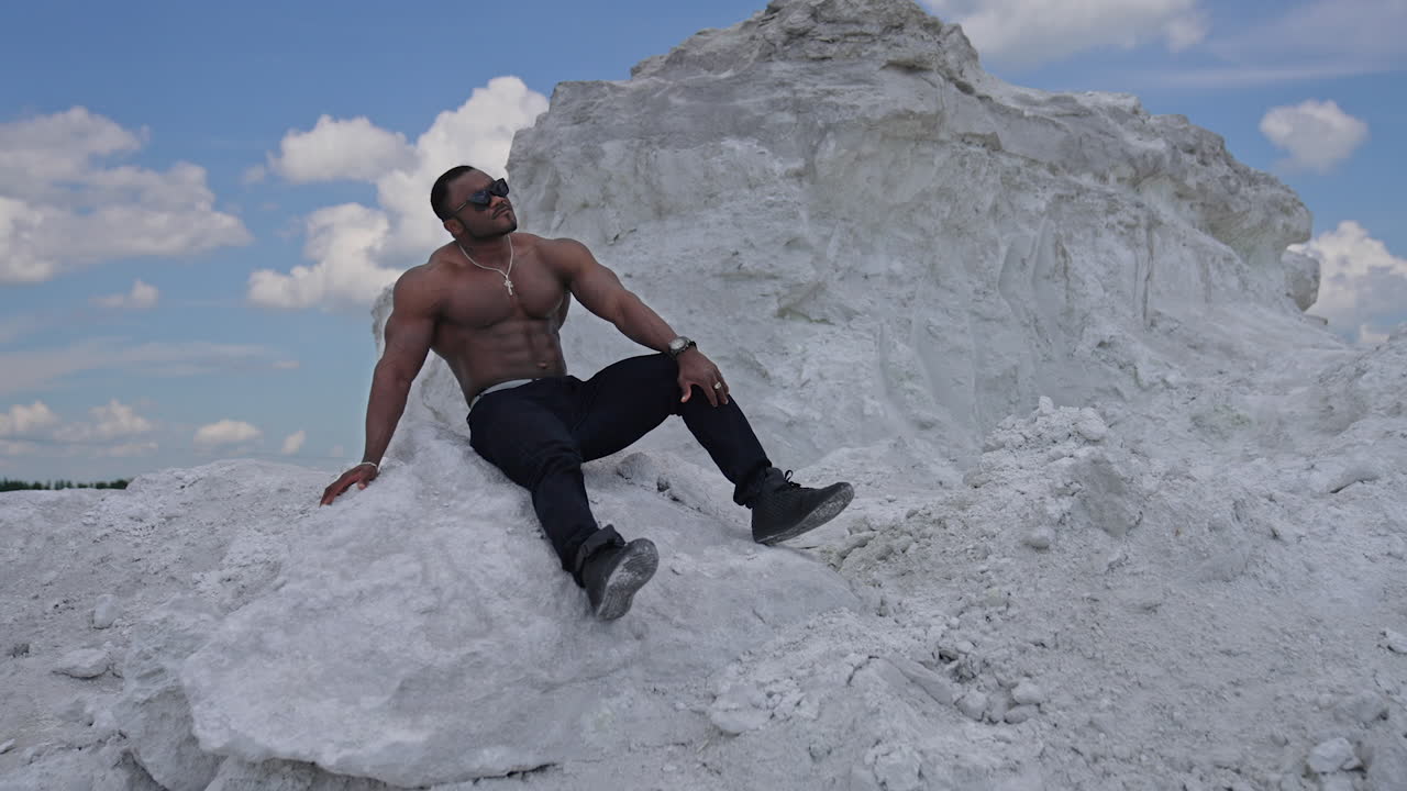 Man with perfect body pumping up muscles and enjoying of the summer. Multiracial man sitting at the sand. Grey rocks background
