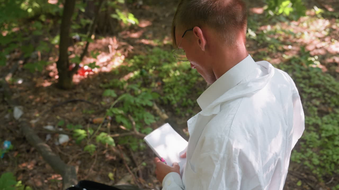 Back view of biologist wearing white lab coat standing near black backpack on old tree stump holding jotter and pen while observing forest environment ready to take field notes in natural setting