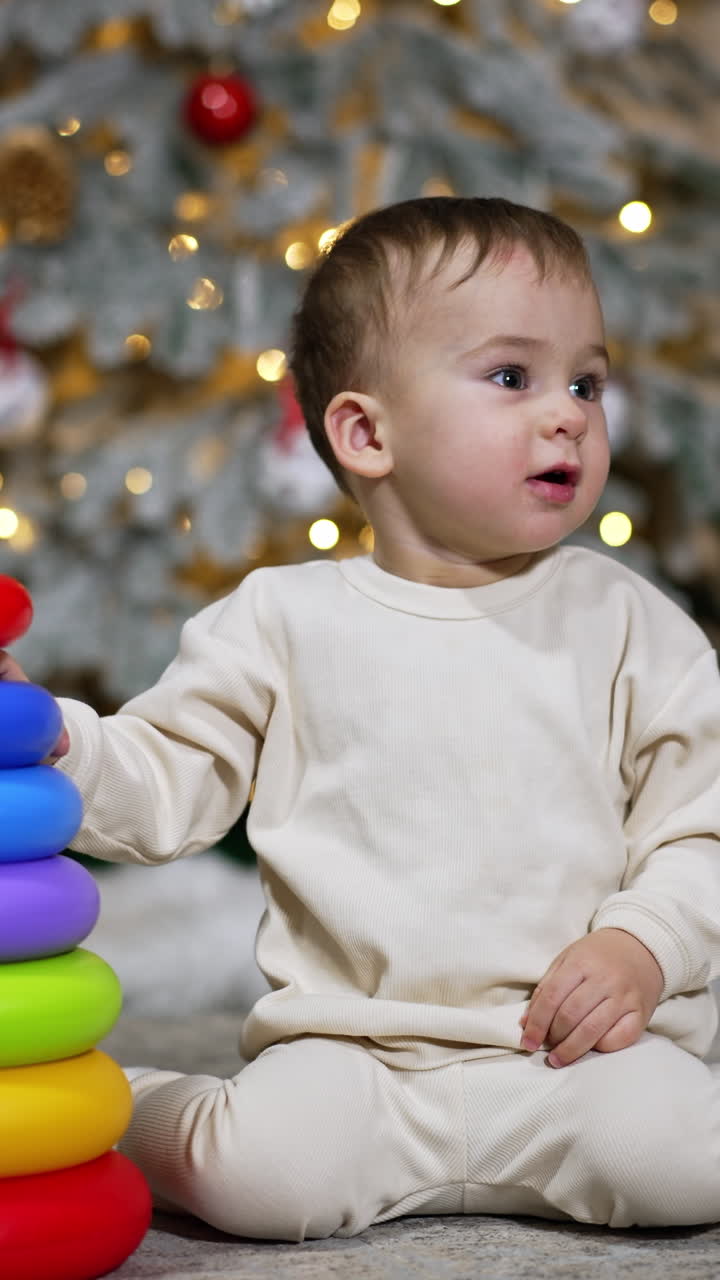 Happy baby boy collecting the pieces of toy pyramid. Toddler is waving hands happily and smiling. Black cat walking by at foreground. Christmas tree at backdrop. Vertical video