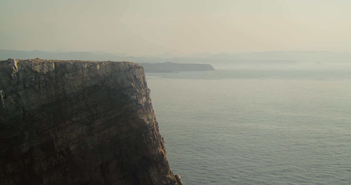 Dramatic aerial footage captures the rugged cliffs of Cabo Peñas on the Asturias coast, bathed in soft, dim sunset light. Wide panoramic view, tranquil Atlantic water, serene natural beauty.