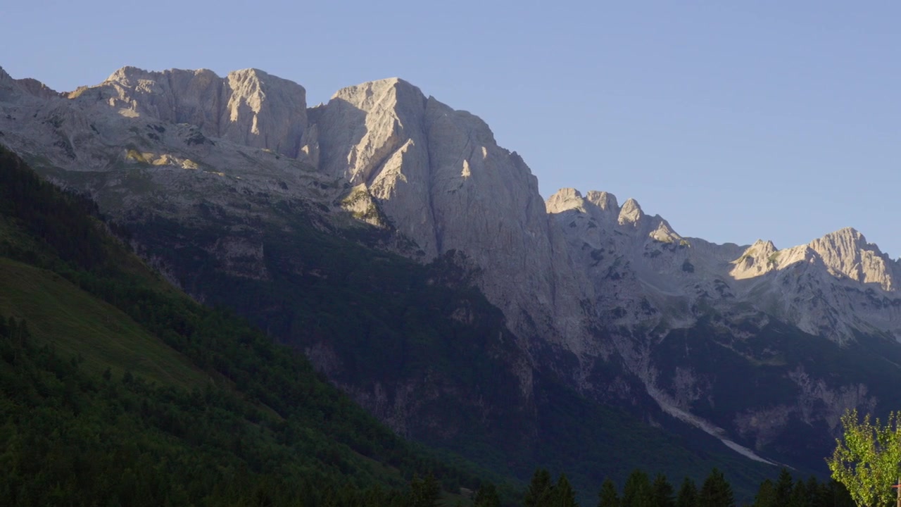 La lumière du soleil illumine progressivement les sommets rocheux et la forêt verdoyante de la vallée de Valbona sous un ciel bleu clair, capturant la lumière du petit matin sur les montagnes albanaises.