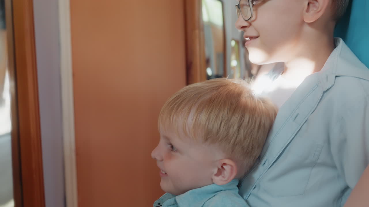 White brothers laugh at mirror, older sibling in glasses comforting younger child with hand on shoulder as sunlight glints on curved glass, candid indoor family moment full of amusement and warmth
