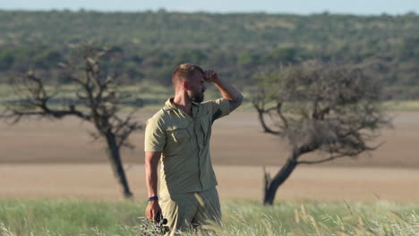 tourist guy standing on grassfield looking through a telescope enjoying the wildlife view in central kalahari game reserve, botswana, southern africa