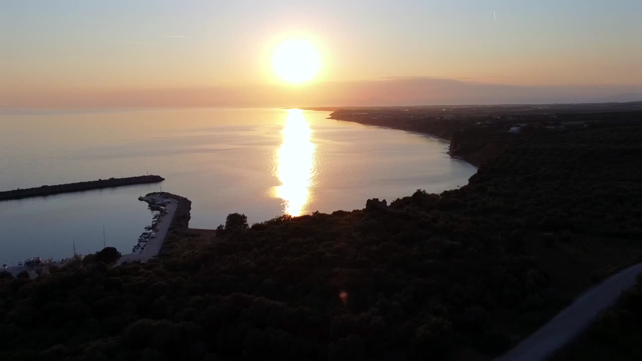 Tomada cinematográfica aérea de drone com recuo revelando o pequeno porto de Agios Charalambos em Maroneia, Grécia. A filmagem transita lentamente das colinas verdes exuberantes para o mar calmo, a praia de areia