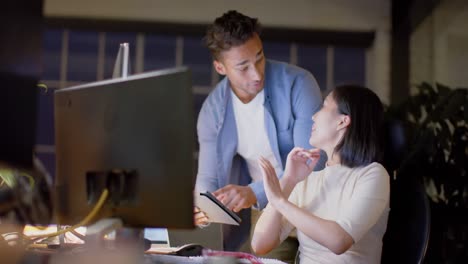 Happy-diverse-male-and-female-colleague-talking-and-using-computer-and-tablet-at-night-in-office