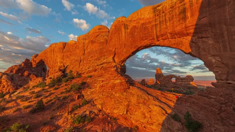 Goldener-Sonnenaufgang-Im-Zeitraffer-Mit-Lichtstrahlen-Auf-Einzigartige-Geologische-Formationen-Und-Fließende-Wolken-An-Den-Fenstern-Und-Dem-Turmbogen-Im-Arches-Nationalpark,-Utah,-Vereinigte-Staaten
