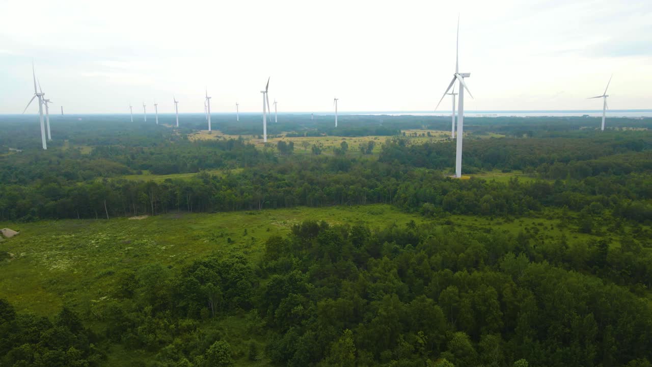 Aerial drone view of large white renewable energy wind turbines spinning and rotating on a cloudy and foggy peninsula during a summer or autumn cloudy day with foret and roads around. Baltic sea also