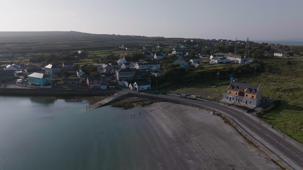 Eine Drohnenaufnahme aus der Vogelperspektive, die das Küstendorf Kilronan auf Inishmore zeigt. Mit Hafen, lokalen Häusern und einem Sandstrand unter klarem Himmel.