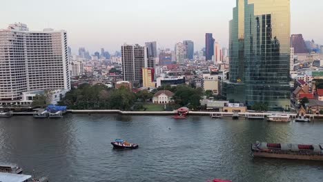 drone video capturing bangkok's urban skyline along a bustling river. high-rise buildings reflect soft dusk light, creating a serene and dynamic cityscape from above