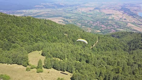 scenic view following paraglider over green trees with valley and town at base of mountain