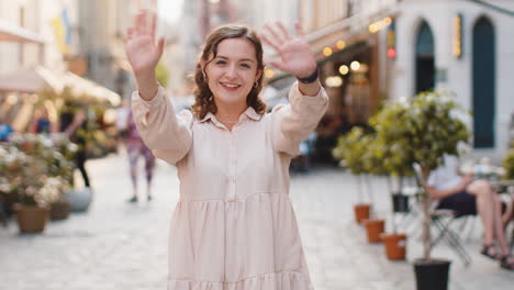 young woman smiling friendly at camera waving hands hello hi greeting or goodbye in city street