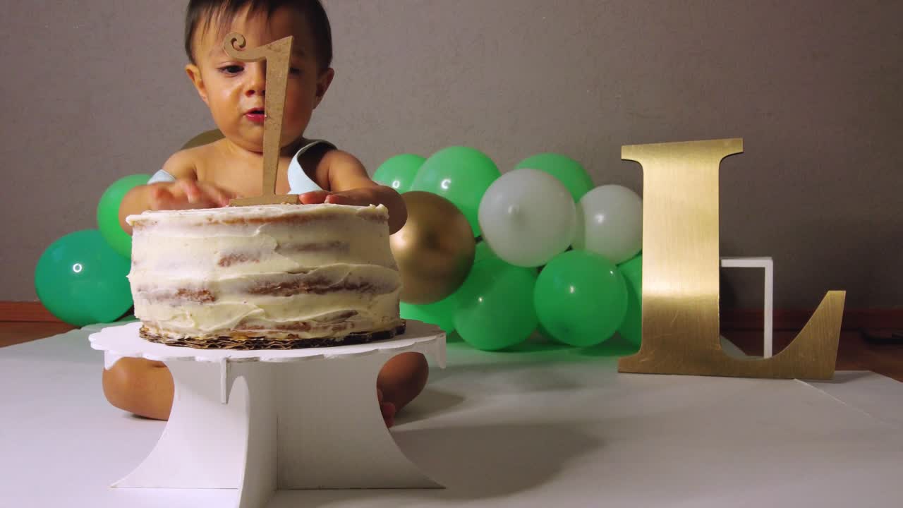 lindo bebé latino celebrando su primer año jugando con su pastel con globos verdes en el fondo