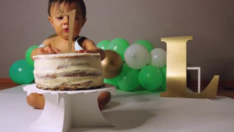 cute latin baby toddler celebrating his 1st year playing with his cake with green balloons in the background