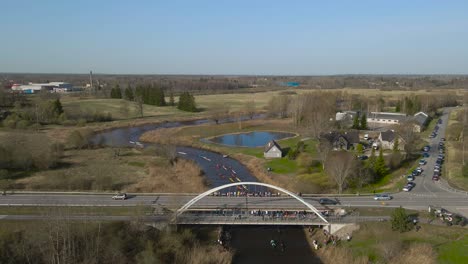aerial drone footage flying backwards and revealing a large kayak and raft marathon on a dark colored river that is flowing under a bridge where spectators and people are watching the race, sunny day.