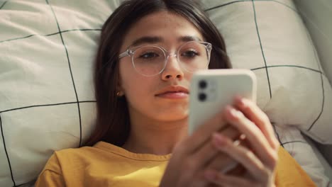 Zoom-in-of-caucasian-teenage-girl-browsing-phone-while-lying-on-bed-on-her-back