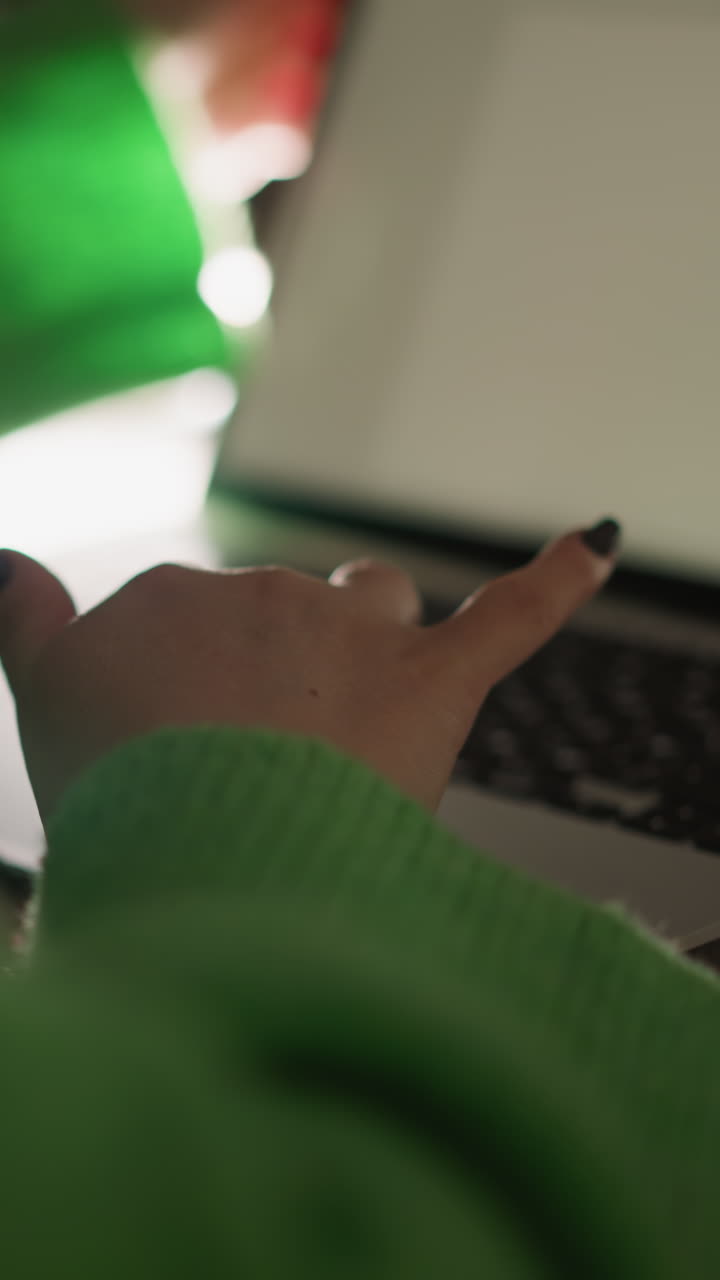 woman in a green sweater working on a laptop while holding a smartphone. Captured with a handheld camera, the scene highlights multitasking, productivity, and modern digital workspace