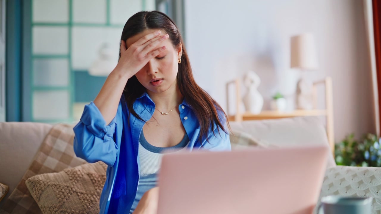 自宅でノートパソコン作業中にストレスを感じ、頭を抱えて困惑するアジア人女性。
