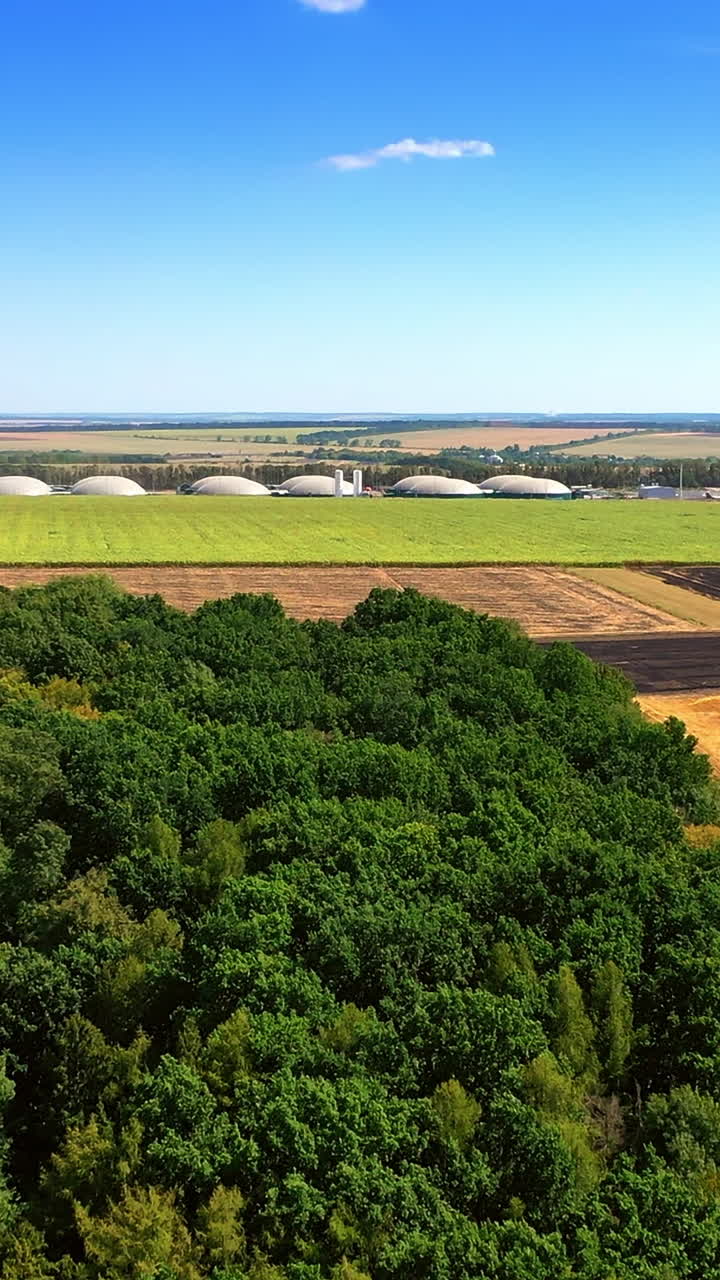 Drone flight over the green woods and fields to the tanks of biogas plant. Sustainable production of green energy. Vertical video