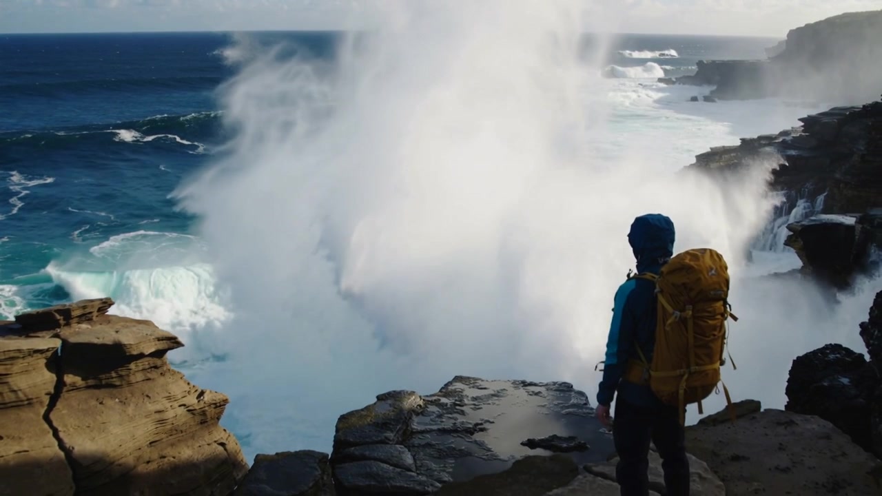 Watching hiker wearing blue jacket, yellow pack facing cliff as huge wave striking reef, copy space