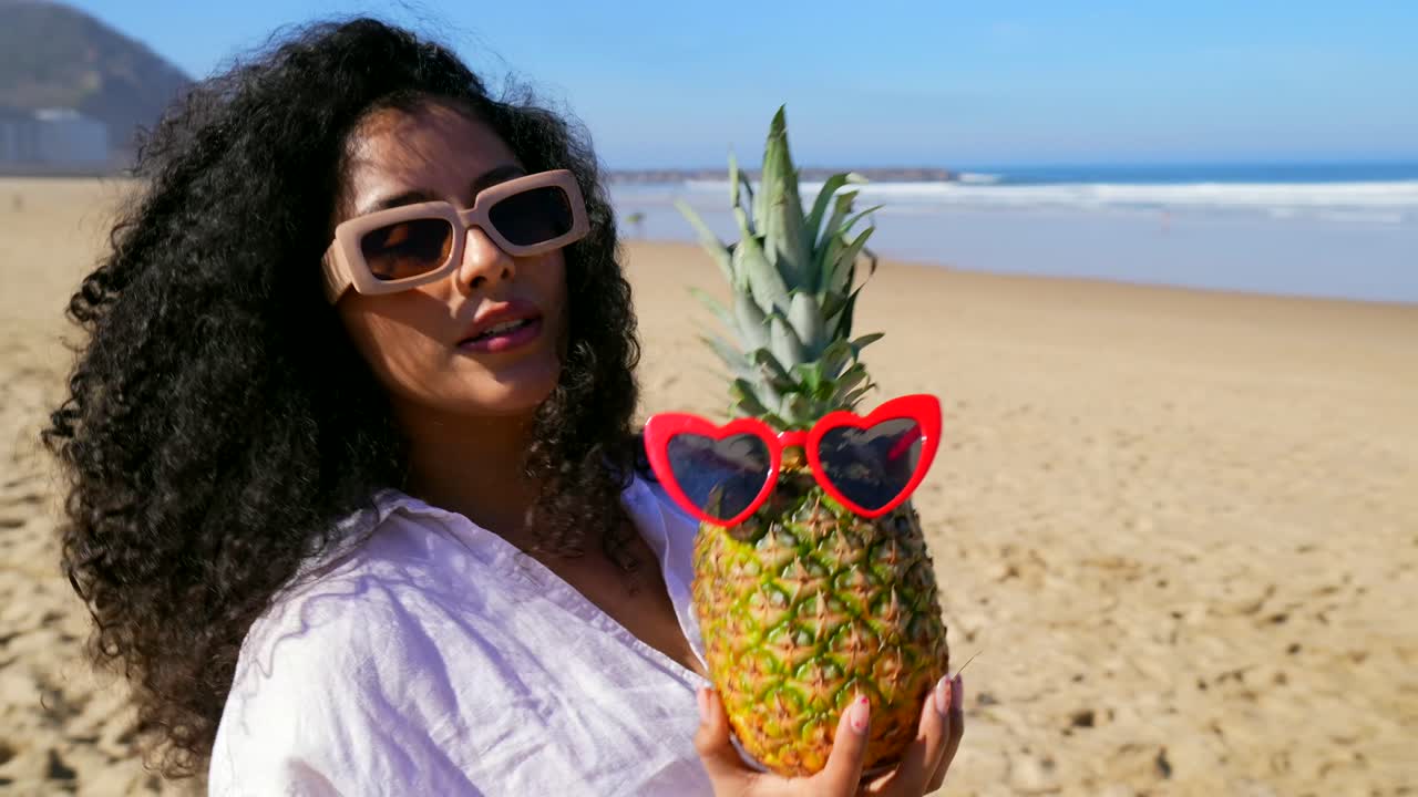 Jovem Mulher Posando com Abacaxi Usando Óculos de Sol em uma Praia Ensolarada