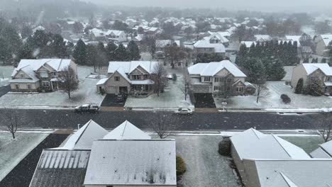 una calle suburbana con casas de dos pisos bajo una ligera nevada, carreteras parcialmente cubiertas y árboles desnudos