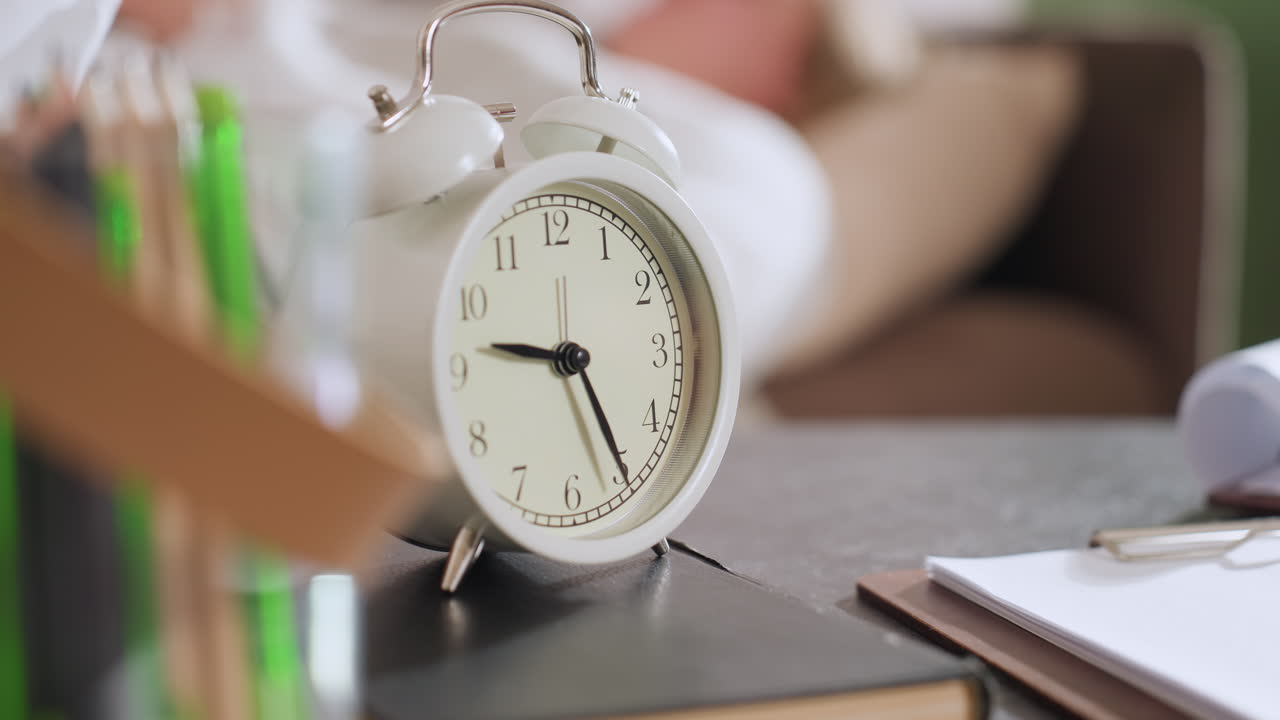 Close up of retro alarm clock on table atop notebook showing time next to clipboard and blurred pens hinting workspace setting conveying focus on schedule timing and organized environment