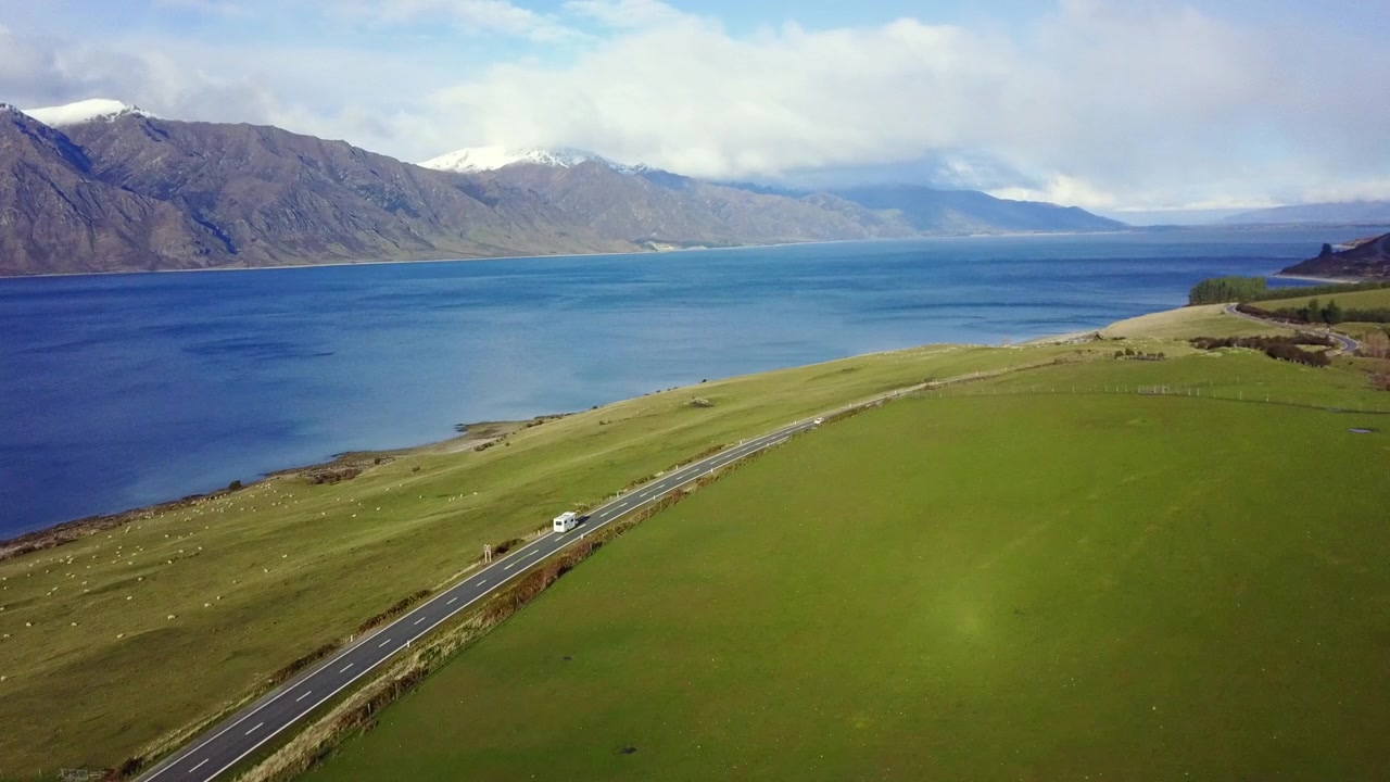 Video aereo di un furgone che guida su una strada costiera con pascoli verdi, oceano blu e montagne innevate