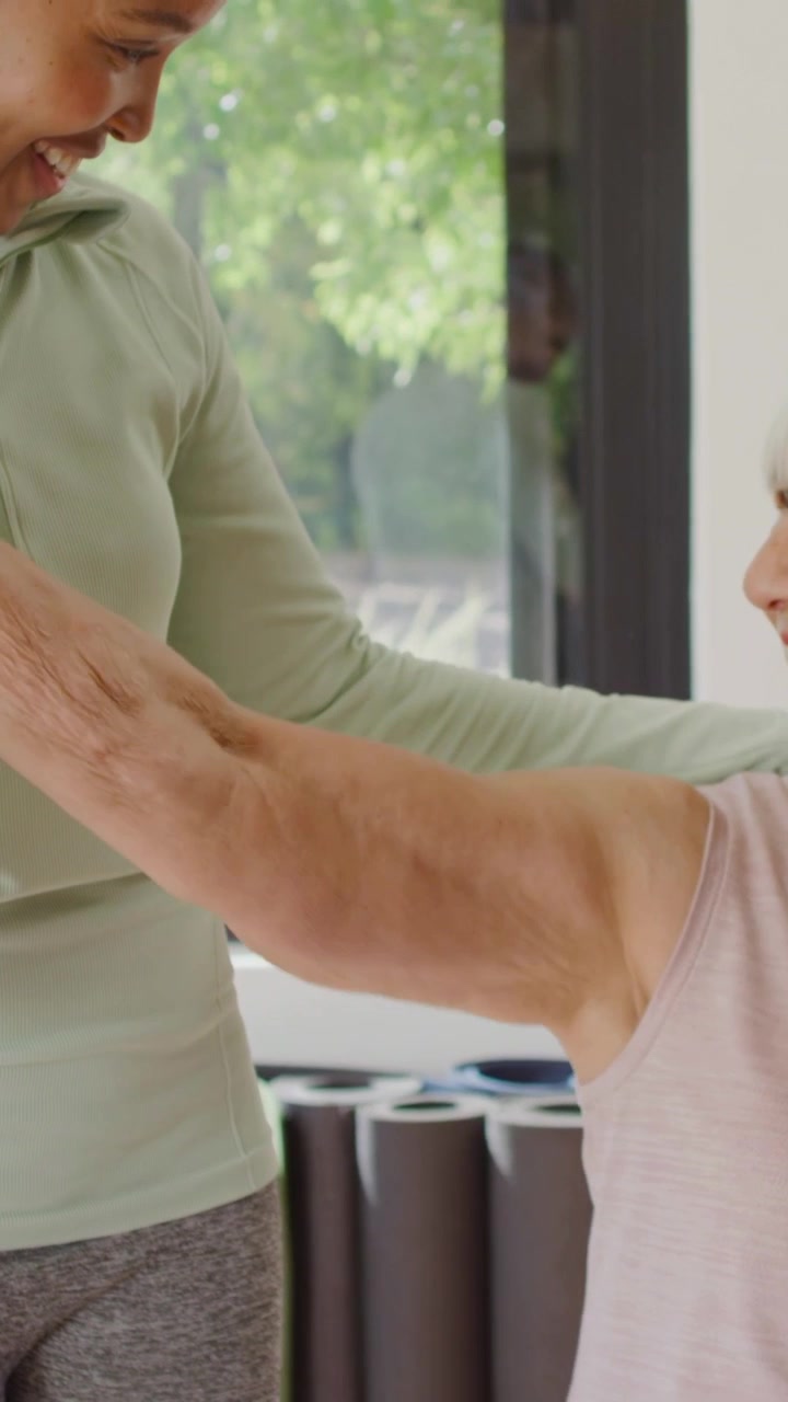 Vertical video: Senior raising arm, trainer guiding arm up in gym with mats pullover aiding stretch