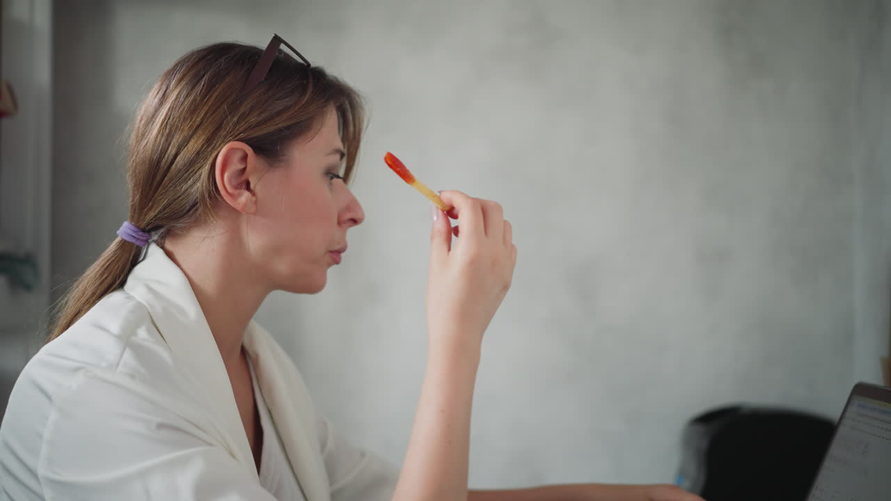 Remote worker dressed in white eating fries dipped in sauce while working on computer stays focused on screen in home office setup with light background