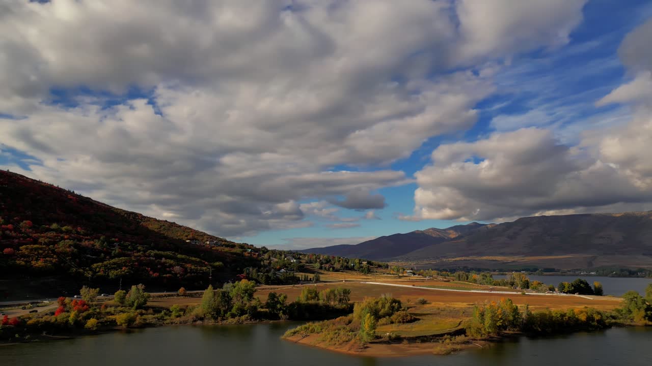 drone movimiento toma de un hermoso lago embalse y vastas montañas plagadas de árboles en el otoño, con naranja, rojo y amarillo árboles y brillante, cielo azul nublado