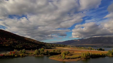 moving drone shot of a beautiful lake reservoir and vast tree plagued mountains in the fall, with orange, red and yellow trees and bright, cloudy blue sky