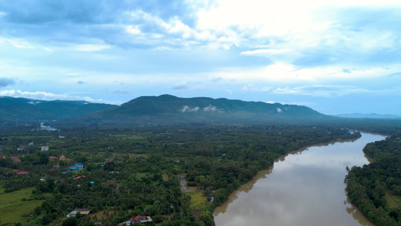 Vue aérienne d’une large rivière traversant des terres agricoles verdoyantes et des collines brumeuses dans la province de Kampot, près de la frontière vietnamienne. Paysage rural paisible avec de vastes paysages champêtres.