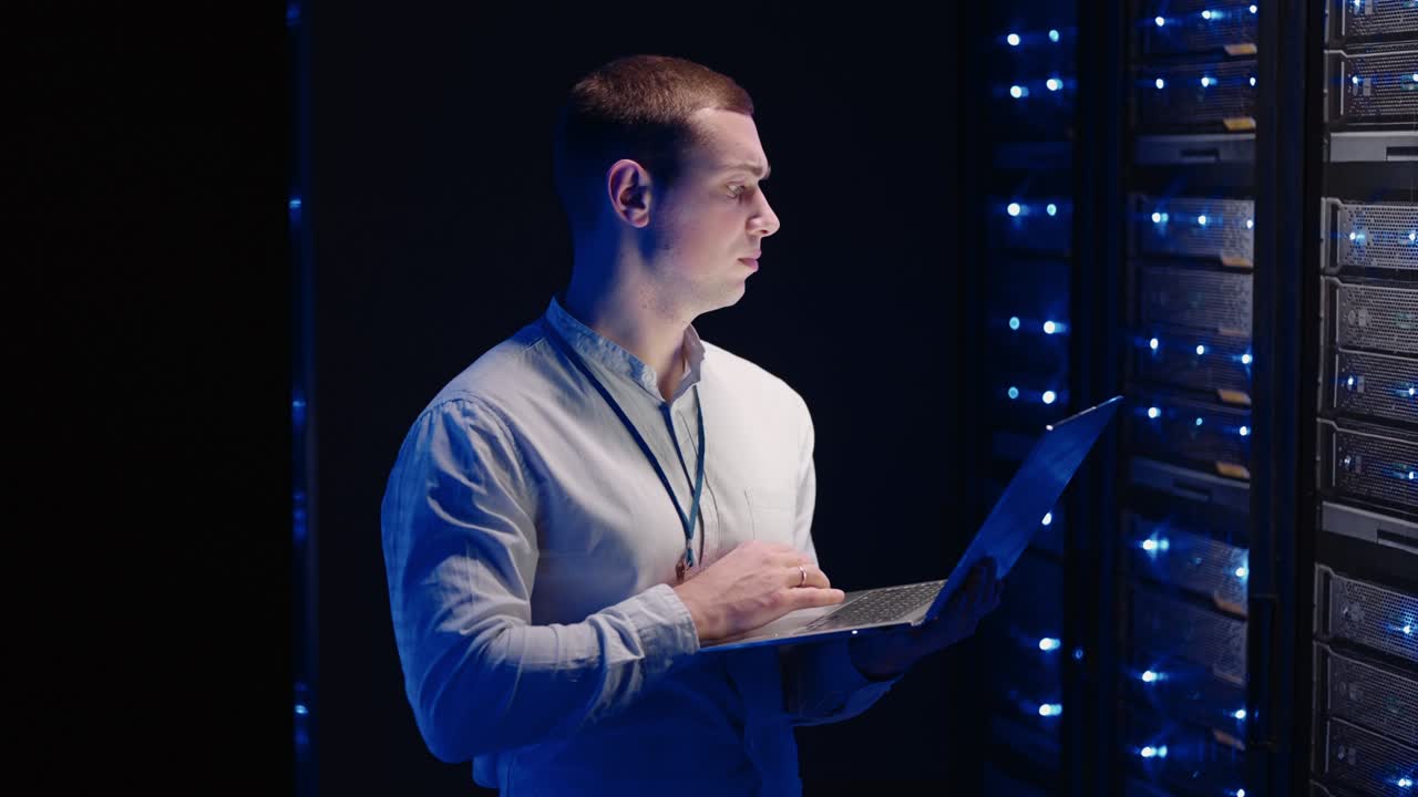 Young man holds device in hand and looks at screen, inspects equipment or hardware rack. Male programmer working with laptop and supporting service while standing in data center spbas.