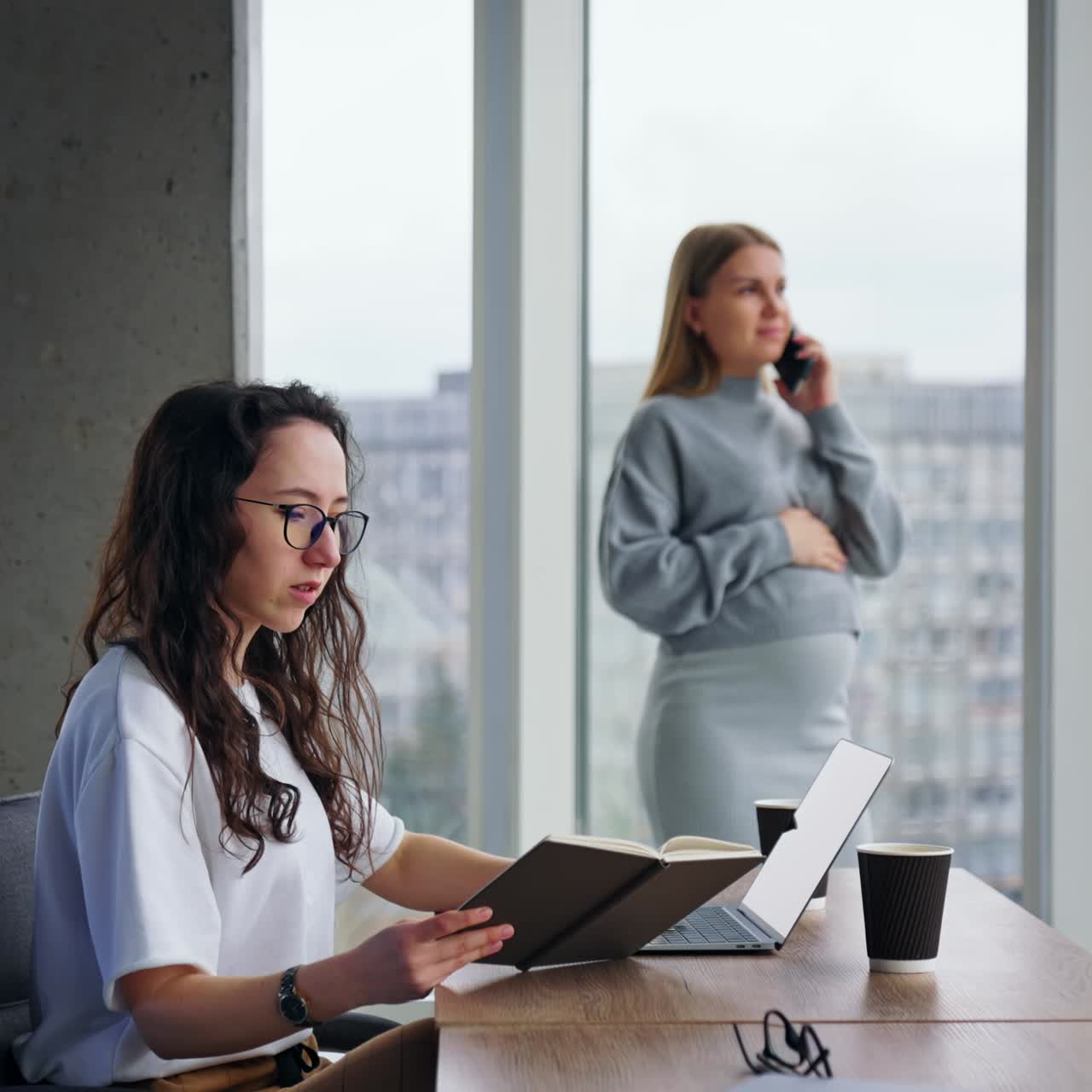 Female with long curly hair types on laptop holding a paper notebook in hand. Blonde pregnant lady stands at window speaking on the phone