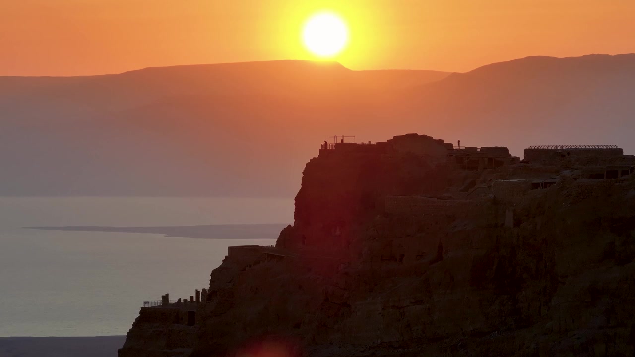 Filmagem cinematográfica com teleobjetiva mostrando o sol brilhante nascendo acima dos cumes das montanhas, projetando um brilho laranja intenso atrás da silhueta escura da antiga fortaleza de Massada, junto ao Mar Morto