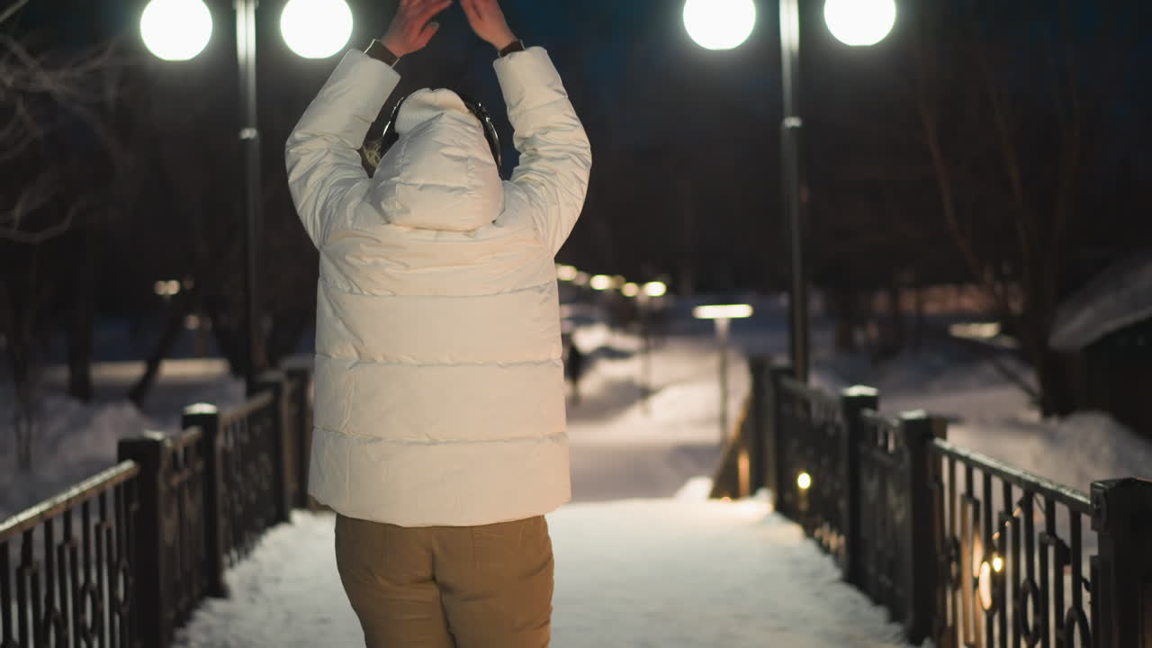 Close up back view of joyful female in white puffer coat with arms raised dancing on snow covered walkway under lampposts at night winter scene expressive movement passionately expressing rhythm