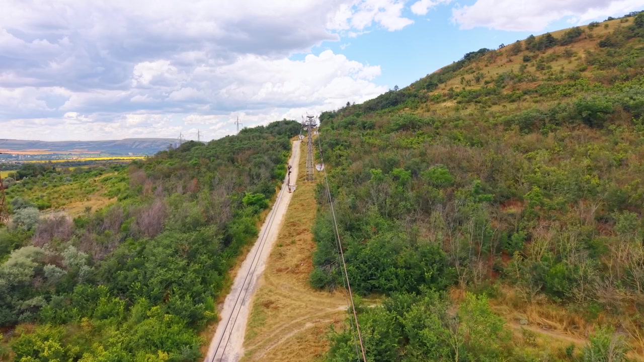 Funivia industriale lungo una collina erbosa sotto un cielo nuvoloso. La vista prospettica mostra una struttura di supporto in acciaio, funi e benne sospese.