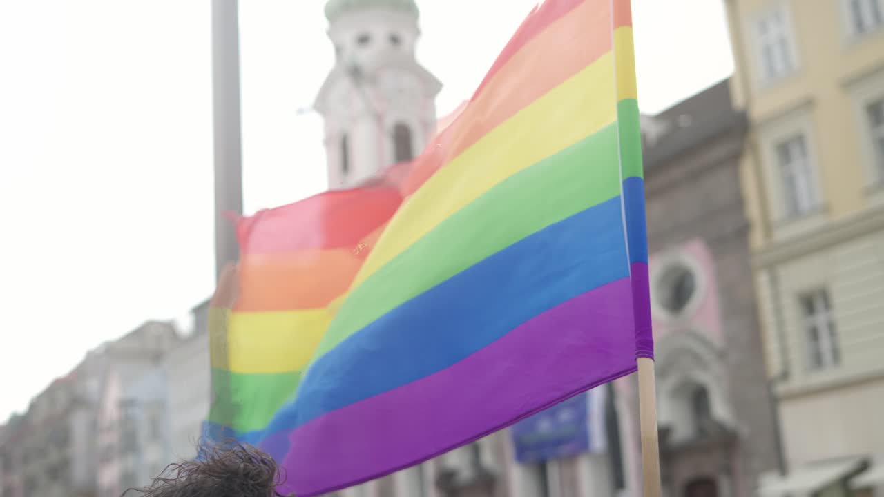 cámara lenta de una bandera lgbtq arcoiris ondeando en el aire durante una manifestación de orgullo en un fondo urbano con casas en el fondo borroso